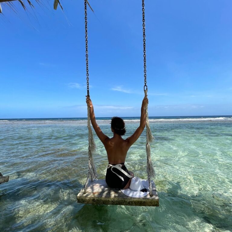 A woman sitting on a swing overlooking the clear turquoise waters of Roatan, Honduras, with a blue sky and ocean in the background.