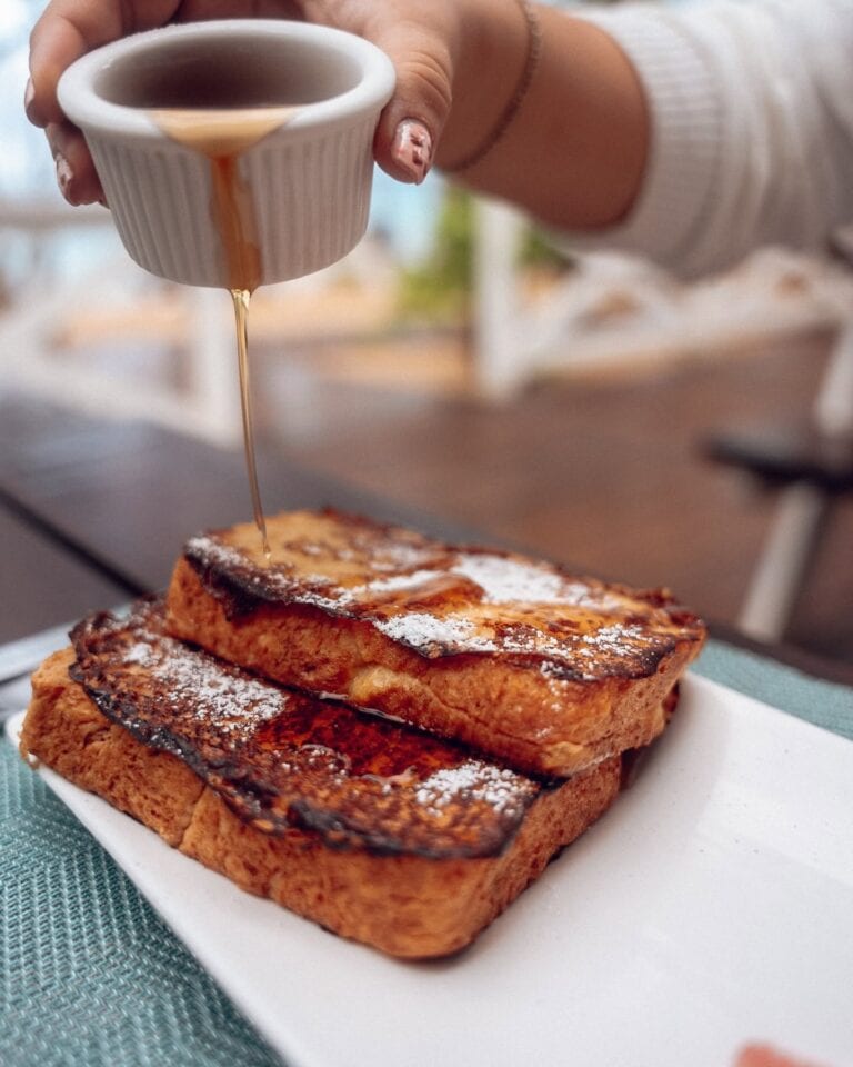 A person pouring syrup over crispy slices of French toast topped with powdered sugar, served on a white plate in Roatan, Honduras.