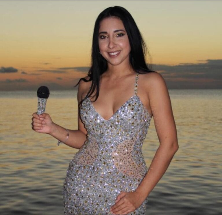 A woman with long dark hair wearing a sparkling silver dress holding a microphone stands on the beach during sunset in Roatan Honduras.