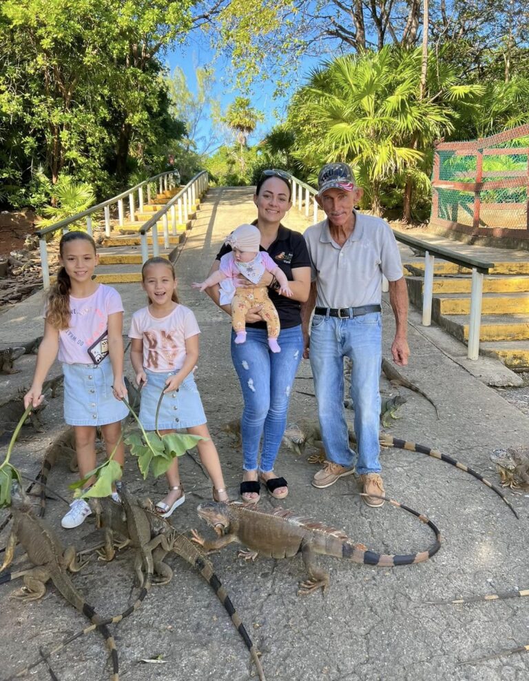 A family of four with two children and an elderly man posing with three large iguanas on a concrete path surrounded by lush tropical greenery in Roatan, Bay Islands, Honduras.