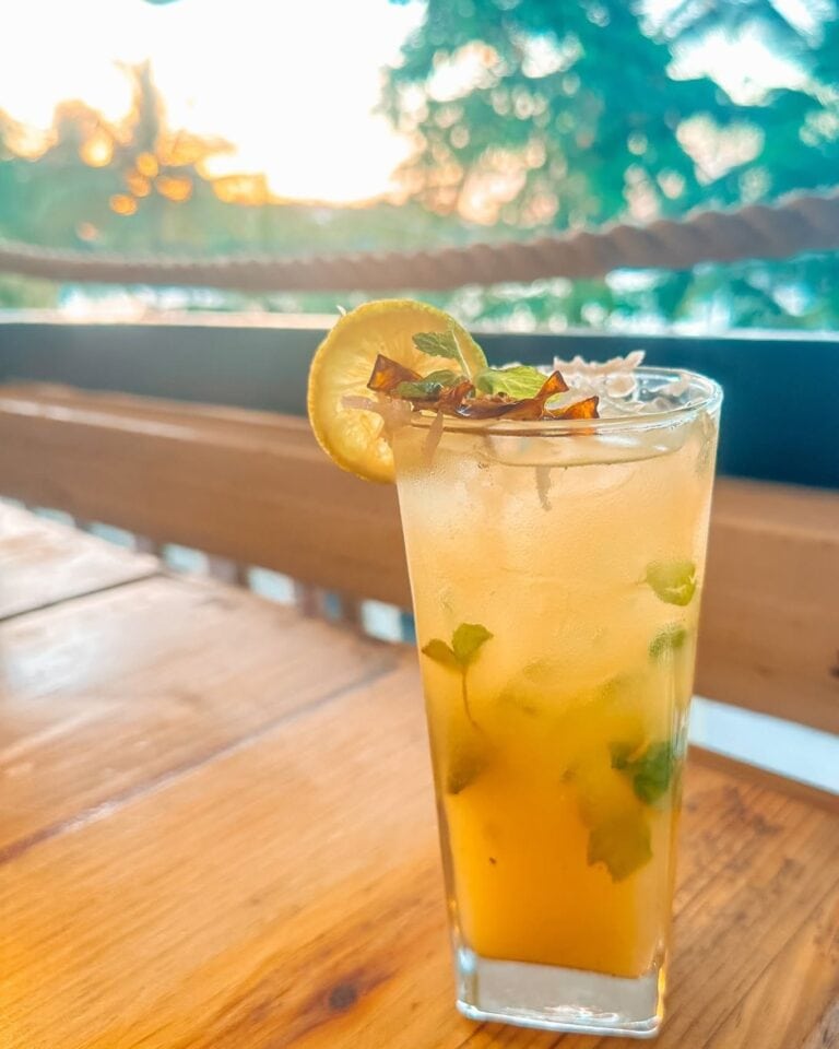 A tall glass of refreshing tropical cocktail garnished with lemon and mint, set on a wooden table with a blurred outdoor background showing trees and sunset in Roatan, Honduras.