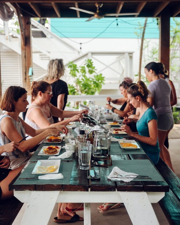 A group of people enjoying a communal meal at an outdoor restaurant in Roatan, Honduras, with a rustic wooden roof and lush green plants in the background.
