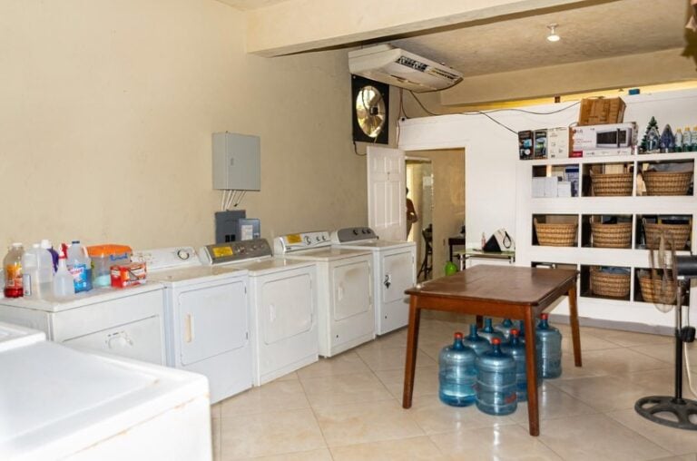 Laundromat with white washing machines and shelves filled with baskets on Roatan, Honduras, featuring a wooden table with large water bottles underneath and cleaning supplies on top.