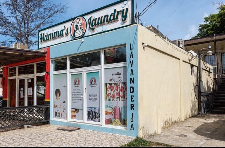 Colorful laundry shop named Mama's Laundry in Roatan, Bay Islands, Honduras, with large glass windows displaying laundry services and colorful towels inside, on a sunny day with a partly cloudy sky.