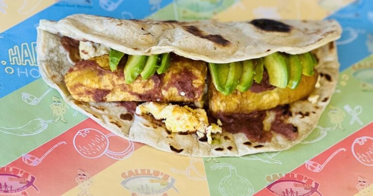 A close-up of a thick, grilled wrap sandwich filled with pieces of grilled chicken, sliced green bell peppers, scrambled eggs, and sauce, resting on colorful printed paper from a food vendor in Roatan, Honduras.