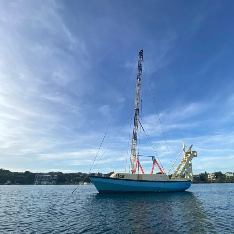 A sailboat named iSoar with a tall mast is anchored on calm waters near Roatan in the Bay Islands of Honduras under a clear blue sky with some wispy clouds.