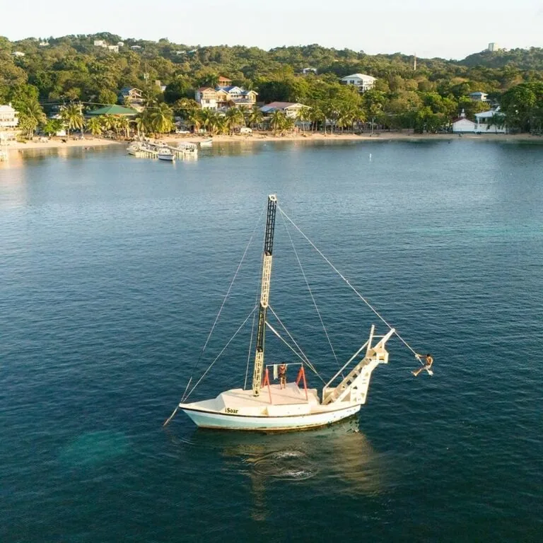A white boat named iSoar with a tall mast and a person swinging from a rope swing attached to the boat's bow on the calm waters of Roatan, Honduras, with a lush shoreline and hillside residential homes in the background.