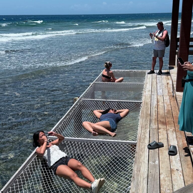 People relaxing on a floating net above the clear waters of Roatan, Honduras, with the ocean and blue sky in the background.