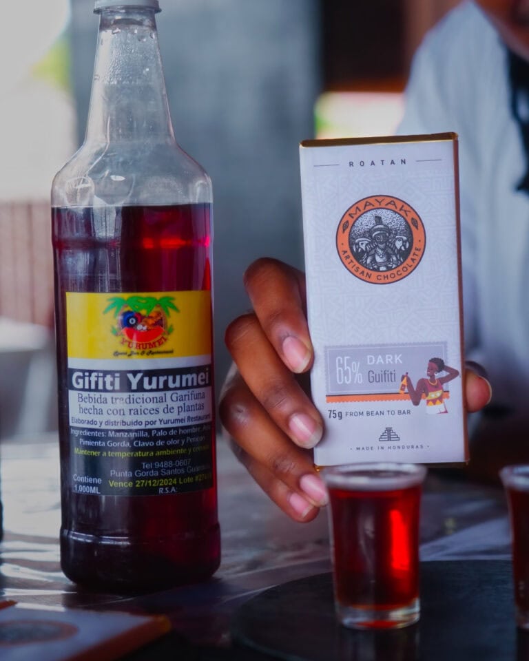 A person holding a bar of dark chocolate from Roatan, Honduras, with a bottle of traditional Yuremei Garifuna medicinal drink and a shot glass filled with red liquid on a wooden table.