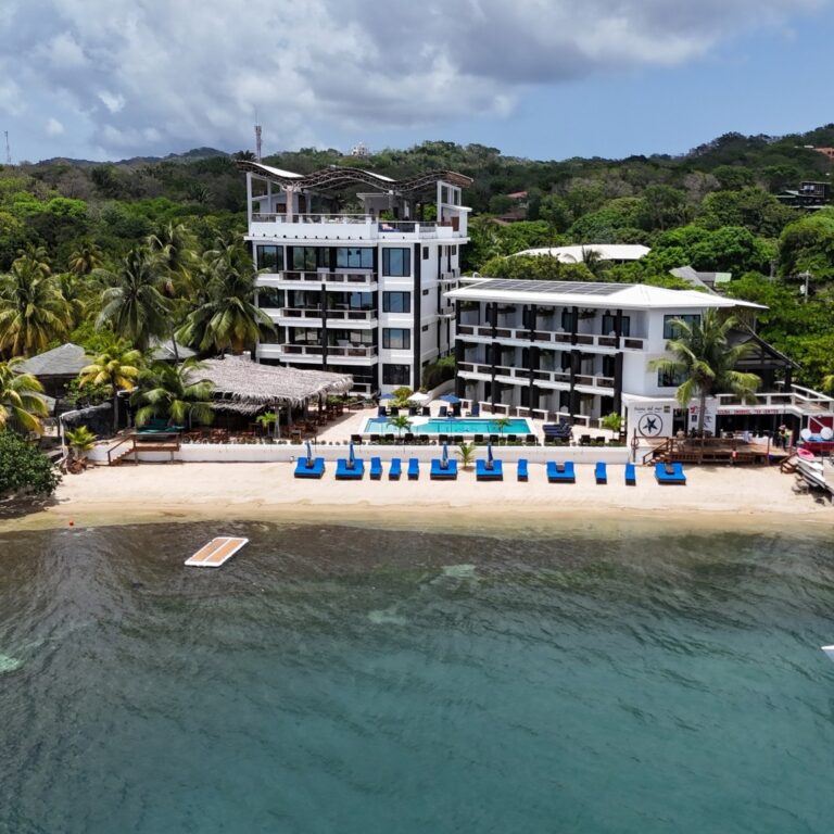 Luxury beachfront hotel with white modern buildings, surrounded by palm trees, and a swimming pool overlooking the Caribbean Sea in Roatan, Honduras.