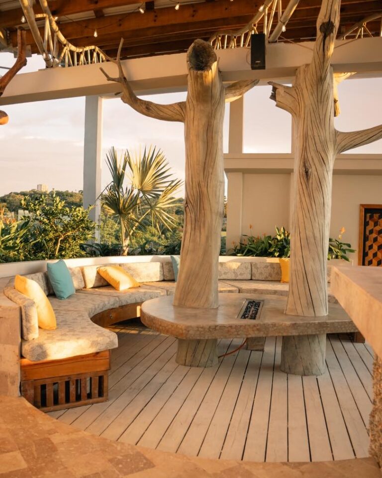 Open-air rooftop lounge in Roatan, Bay Islands, Honduras featuring a circular wooden bench with yellow and turquoise cushions, surrounded by tropical plants and large tree-shaped wooden columns supporting the overhead structure during sunset.
