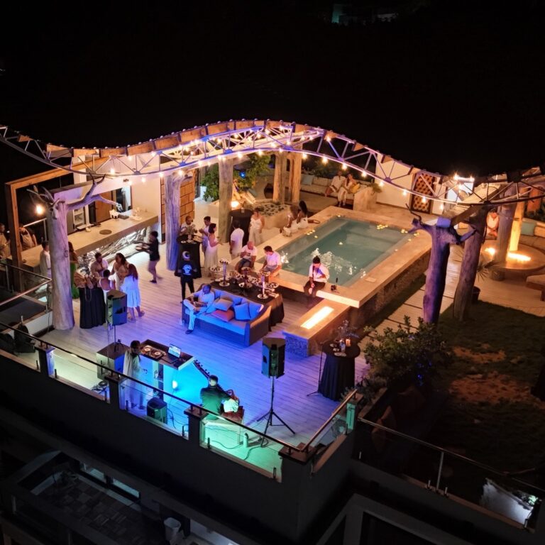 Nighttime view of an outdoor pool party on Roatan, Bay Islands, Honduras, featuring a lit hot tub, DJ, and people socializing in a tropical resort setting.