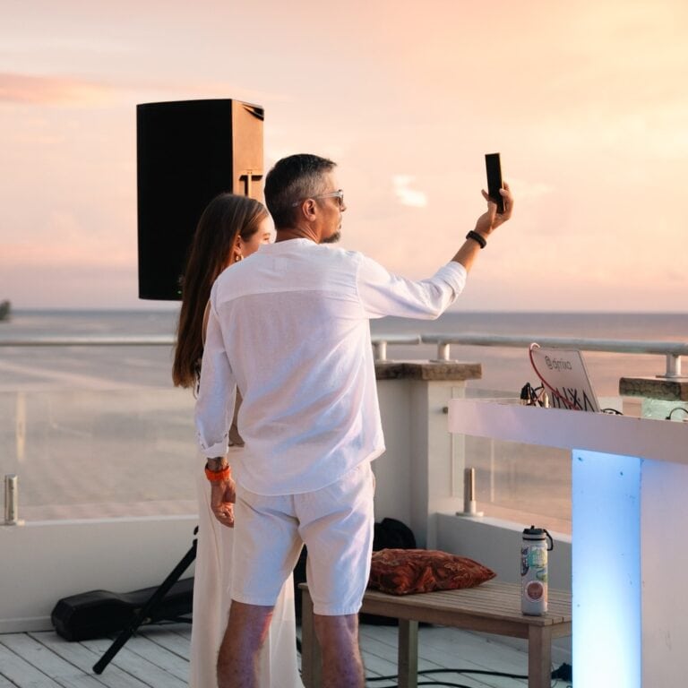 A man and woman taking a selfie on a rooftop terrace overlooking the beach at sunset in Roatan, Bay Islands, Honduras.