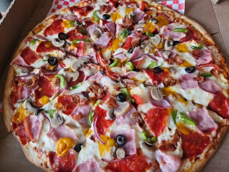 A freshly baked pizza with toppings including ham, mushrooms, green bell peppers, black olives, and melted cheese on a wooden table in Roatan, Honduras.