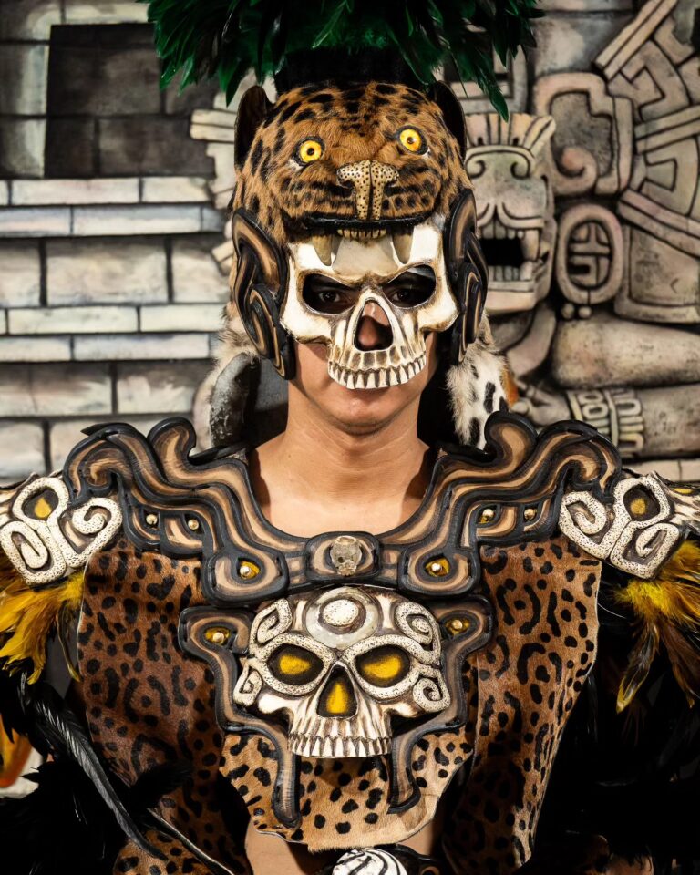 Person dressed in traditional Mesoamerican costume with skull and jaguar motifs, wearing a headdress resembling a jaguar head with yellow eyes in Roatan, Honduras.