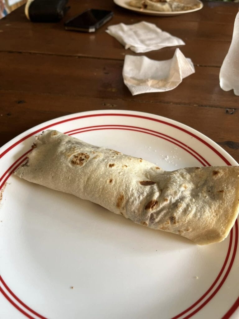 A close-up of a freshly made burrito on a white plate with red stripes, placed on a wooden table in Roatan, Honduras, with condiment packets and a water glass nearby.