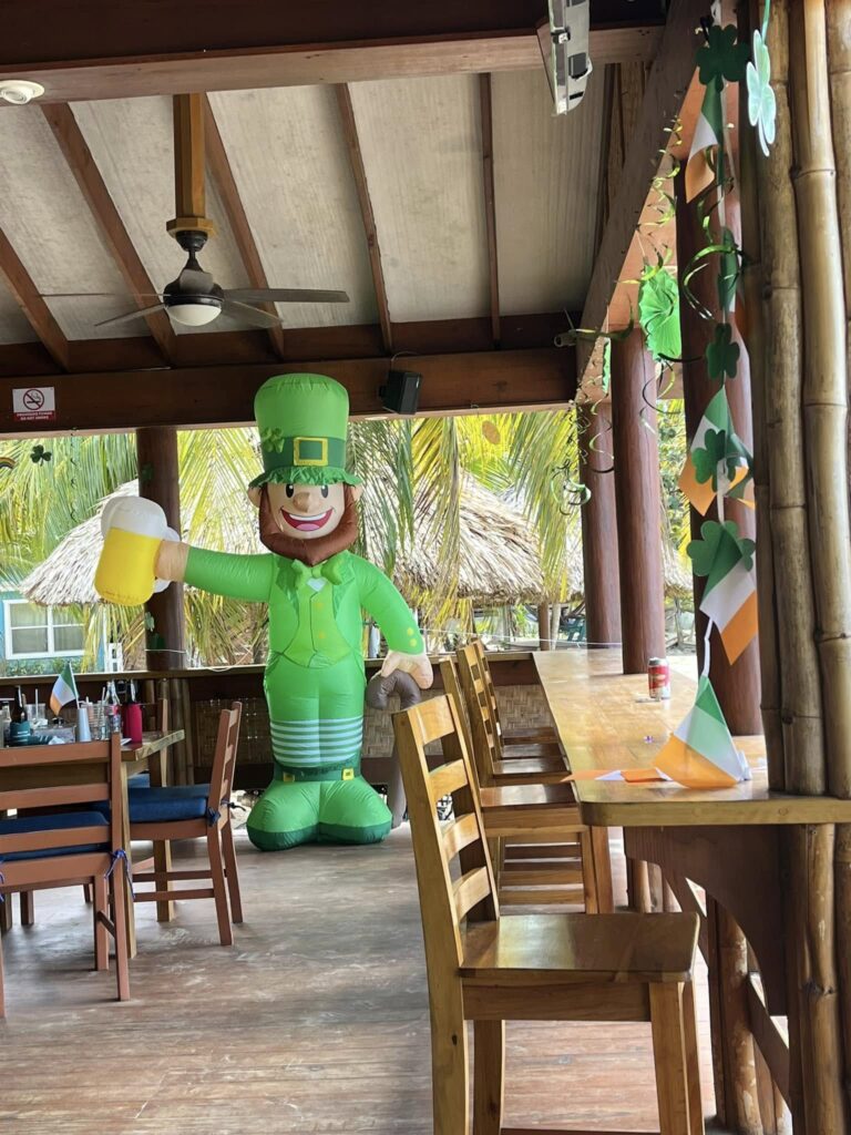 Decorated outdoor dining area in Roatan, Honduras with a St. Patrick's Day theme including shamrock garlands and a large inflatable leprechaun figure holding a beer glass.