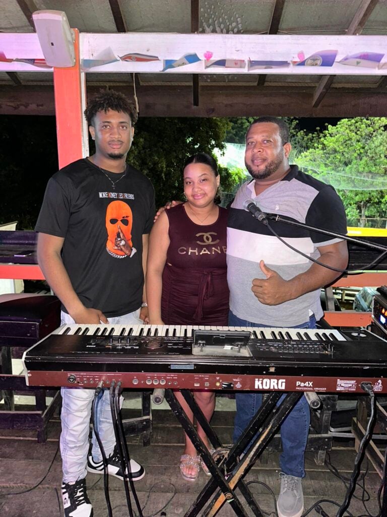 Three musicians perform together under a wooden structure on Roatan, Bay Islands, Honduras, with musical keyboards and a microphone visible as they entertain local guests.