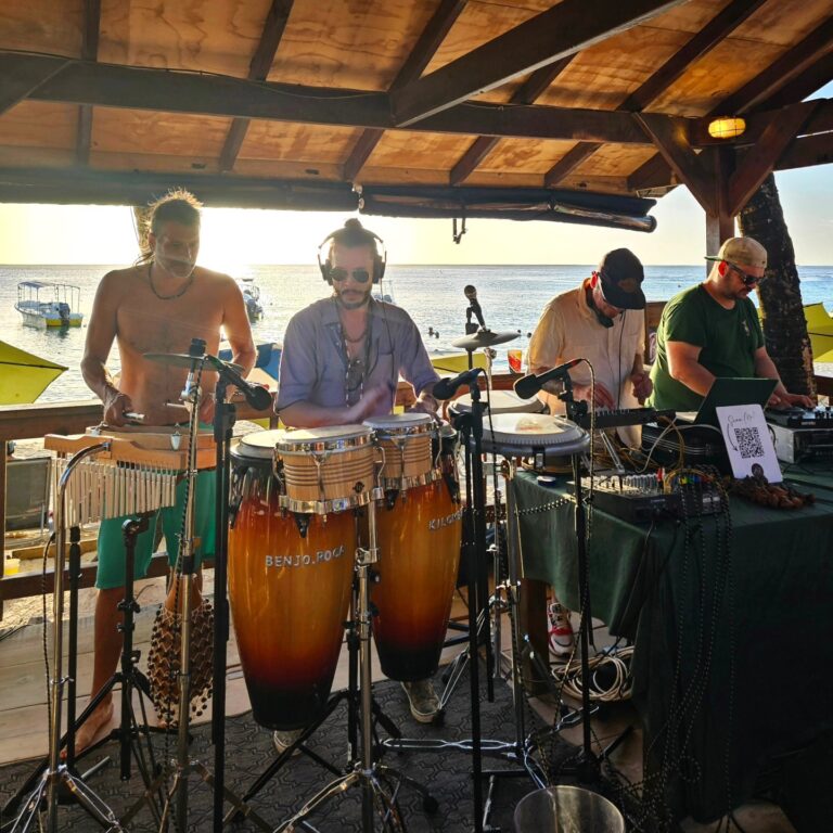 A live band performs on a wooden beachside pavilion in Roatan, Honduras, with the ocean and boats visible in the background during sunset.