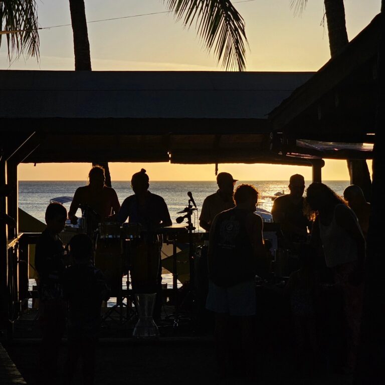 Silhouettes of people enjoying live music on a deck overlooking the ocean at sunset in Roatan, Bay Islands, Honduras, with palm trees in the background.