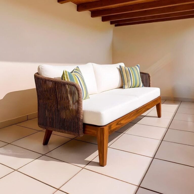 A white sofa with wooden legs and woven wicker sides, decorated with striped cushions in shades of green, blue, and beige, placed in a room with beige tiled flooring and a wooden-beamed ceiling in Roatan, Honduras.