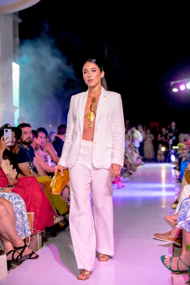 A woman models a white pantsuit with a deep neckline and large gold jewelry on the runway during a fashion show in Roatan, Honduras, with an audience watching.