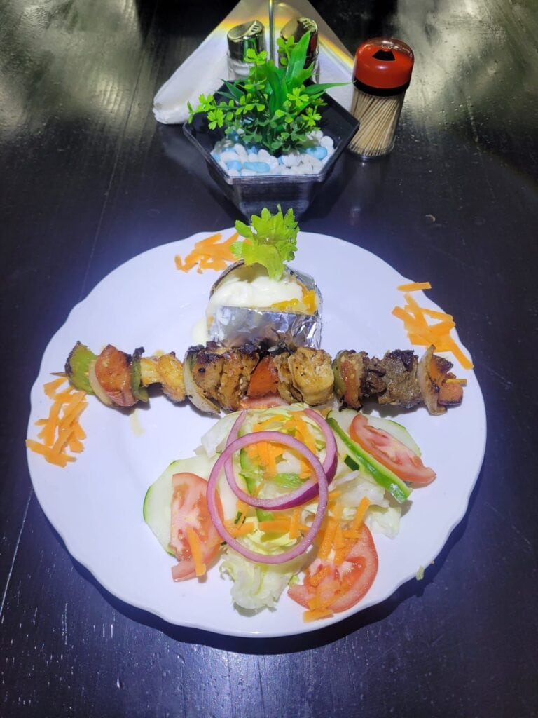 A plate of grilled meat and vegetables served with fresh salad and a baked potato on a white dish, set on a dark wooden table in Roatan, Honduras.
