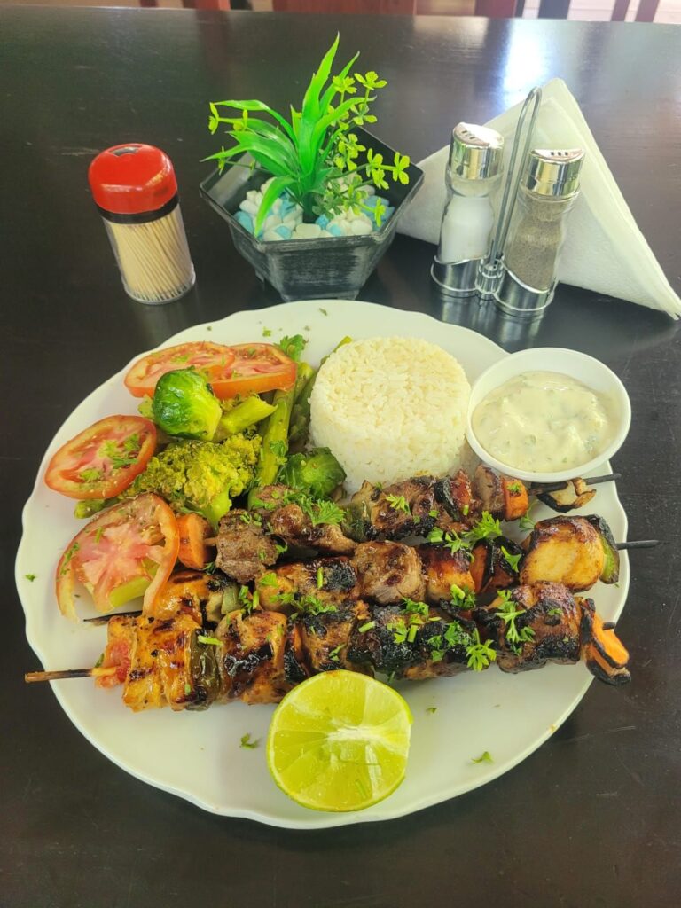 A plate of grilled meat skewers served with white rice, fresh tomatoes, broccoli, and a side of creamy sauce, garnished with lime wedges, on a dark table in Roatan, Honduras.