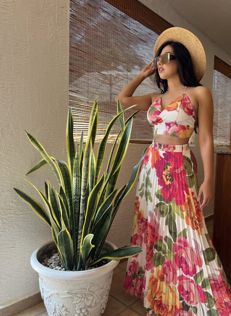 A woman wearing a floral dress, sunglasses, and a straw hat standing indoors next to a large potted snake plant in Roatan, Honduras, with wooden blinds in the background.