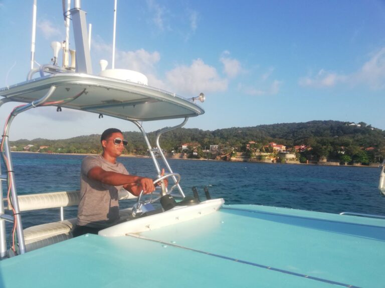 A man wearing sunglasses operates a boat on the clear waters near Roatan, Honduras, with a scenic coastline, houses, and lush green hills in the background.