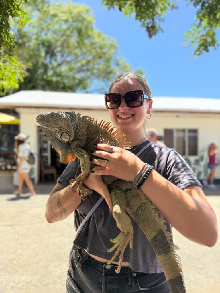 A young woman with sunglasses smiling while holding a large iguana in front of a building on Roatan, Honduras under a clear blue sky.