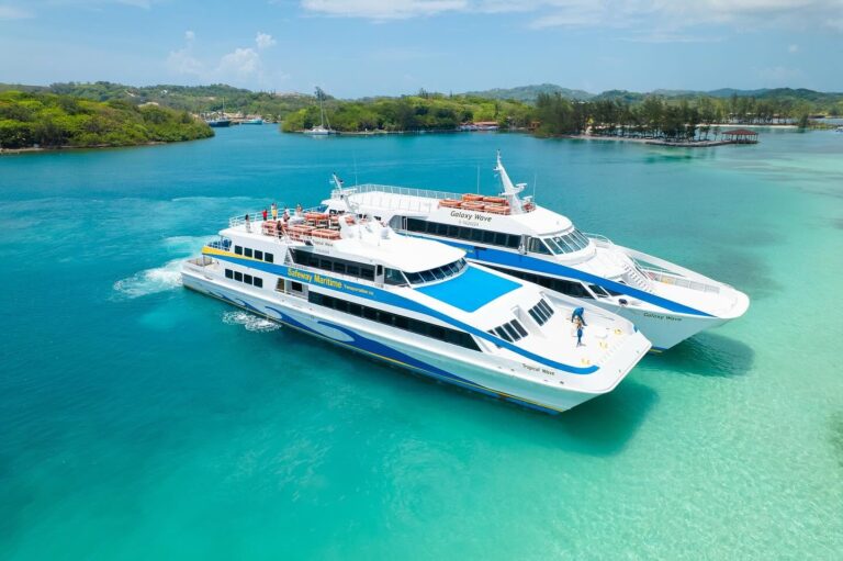 Two large white and blue passenger ferries named Galaxy Wave and Tropical Wave are sailing through the clear turquoise waters of Roatan, Honduras, near a lush green island with trees and some small structures.