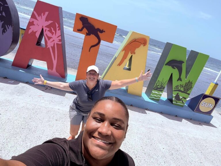 Two smiling visitors in casual clothing pose in front of colorful Roatan and Honduras-themed signs by the beach, with the ocean and clear sky in the background.