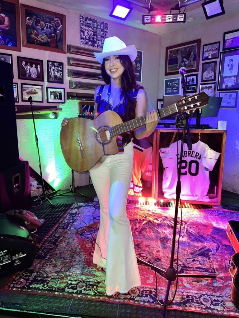 A young woman wearing a white cowboy hat and white pants performs with an acoustic guitar in a cozy bar decorated with framed photographs on the walls in Roatan, Honduras.