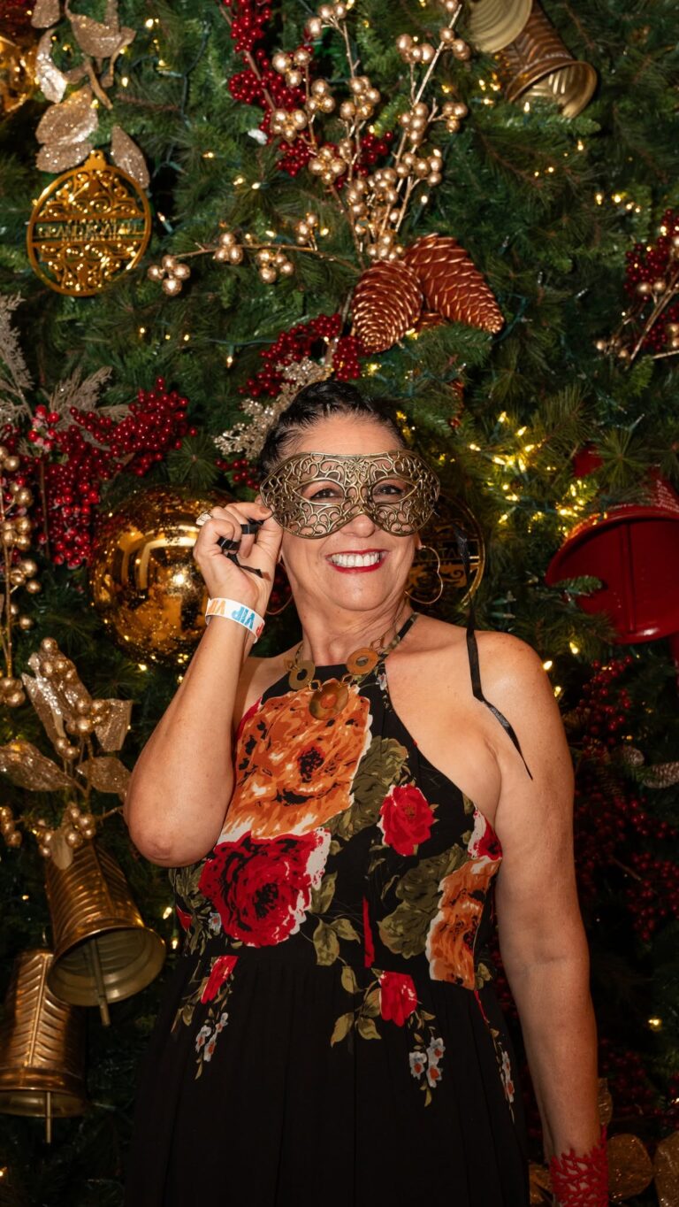 A woman with short dark hair and a floral dress smiling while holding a decorative gold mask in front of a large decorated Christmas tree on Roatan, Bay Islands, Honduras. The tree is adorned with gold, red, and glittering ornaments, pinecones, and warm string lights highlighting the festive setting.
