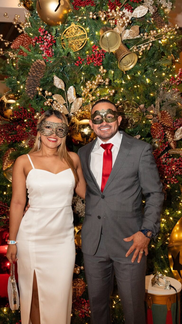 A smiling couple dressed in formal attire wearing masquerade masks stands in front of a lavish Christmas tree decorated with gold and red ornaments, pinecones, and glittering leaves in Roatan, Honduras.