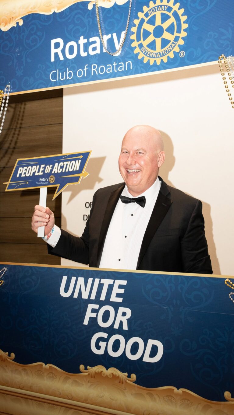 Bald man in a tuxedo holding a sign that reads 'People of Action' at a Rotary event in Roatan, Honduras, with a Rotary International banner in the background.