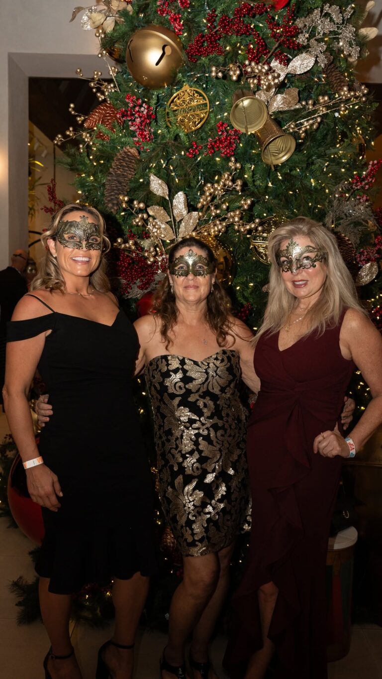 Three women wearing ornate masks stand in front of a decorated Christmas tree in Roatan, Honduras, celebrating the holiday season with festive attire and joyful expressions.