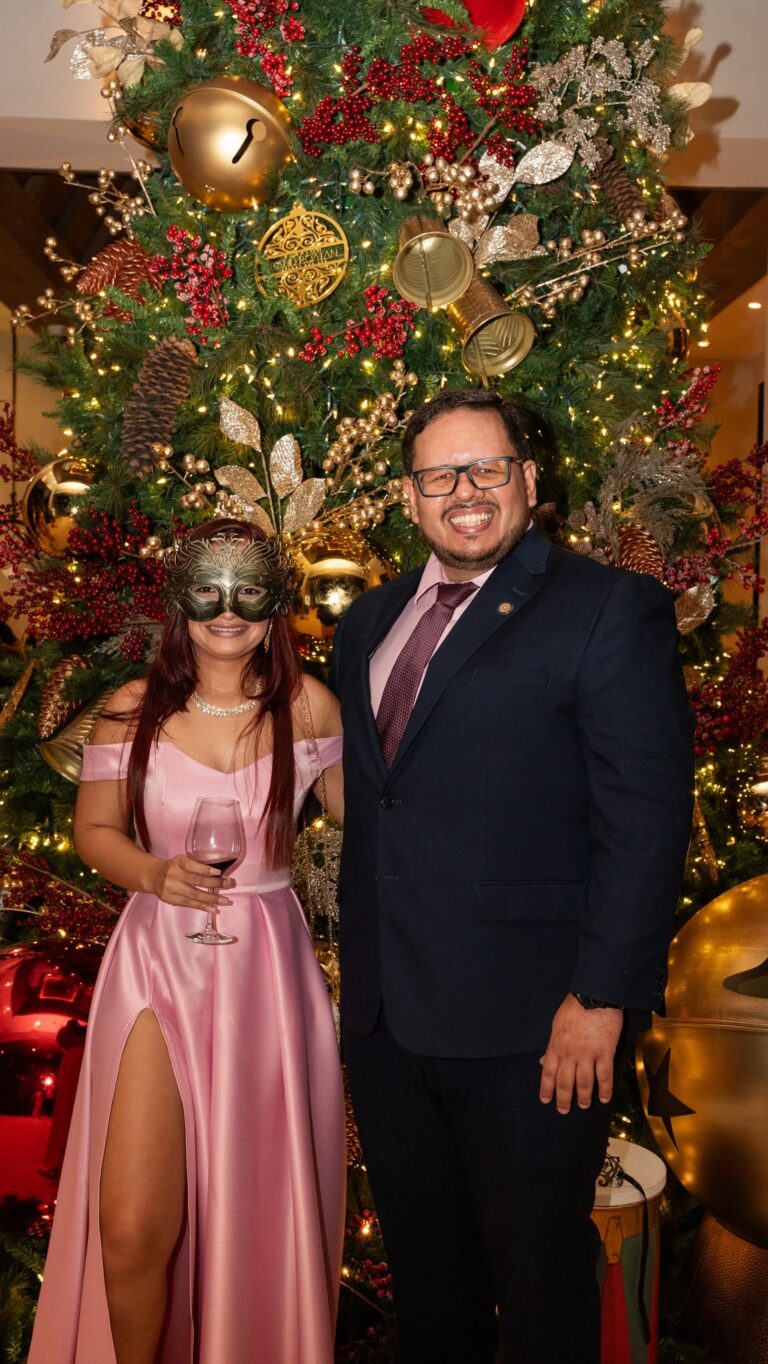 Two people dressed festively stand in front of a decorated Christmas tree in Honduras, celebrating the holiday season on Roatan Island. The woman wears a pink dress with a high slit and a mask, holding a glass of red wine, while the man in a suit smiles beside her.