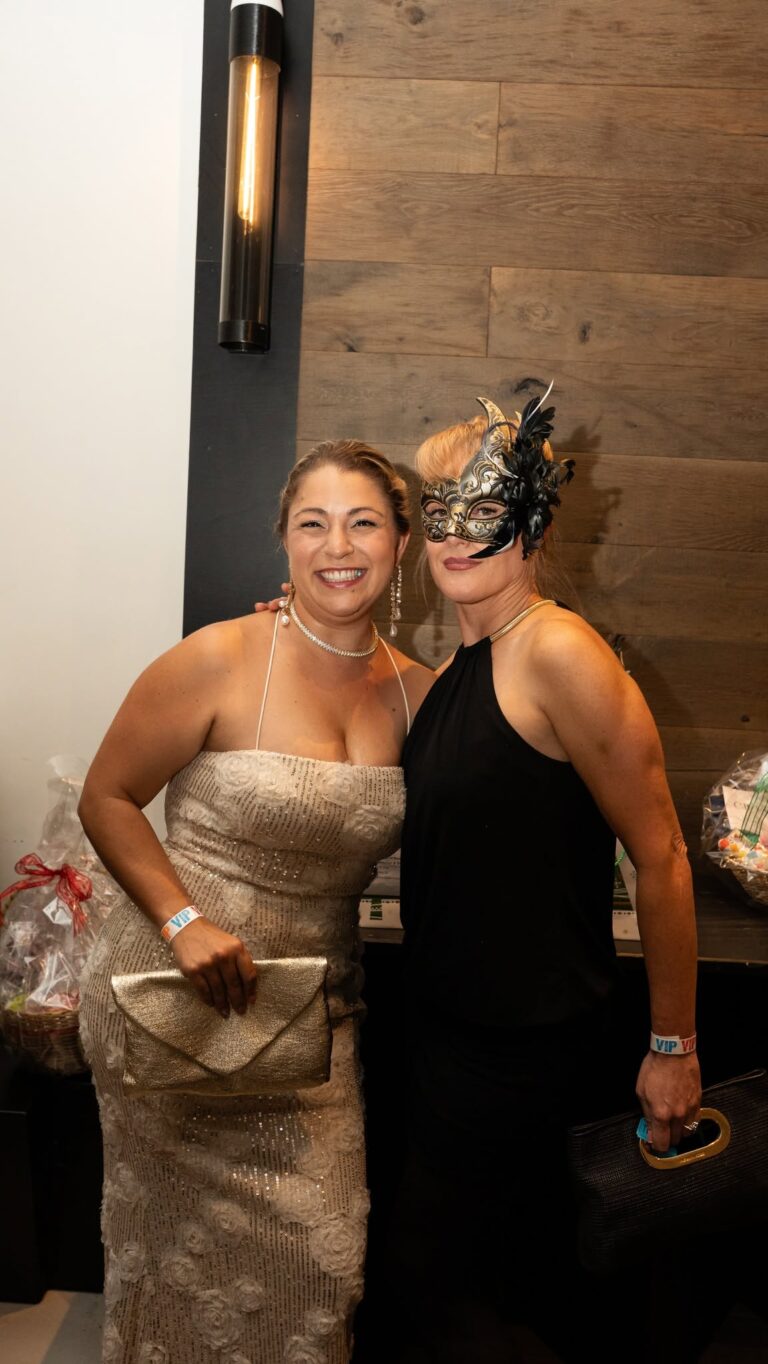 Two women dressed in elegant attire pose together at a social event in Roatan, Honduras, with a wooden wall and lighting fixture in the background.
