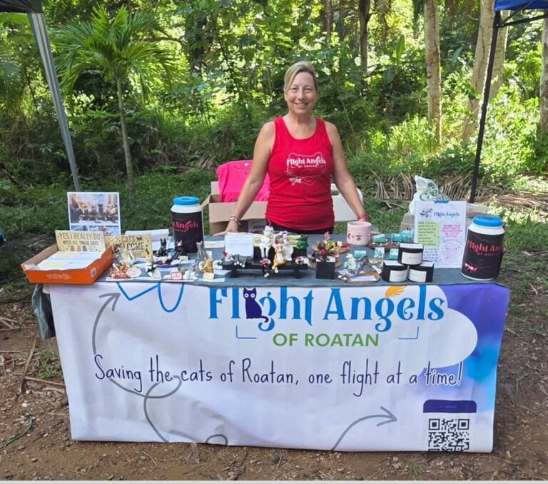 A woman standing behind a table filled with cat rescue merchandise at an outdoor event in Roatan, Honduras, surrounded by lush green trees and forest.