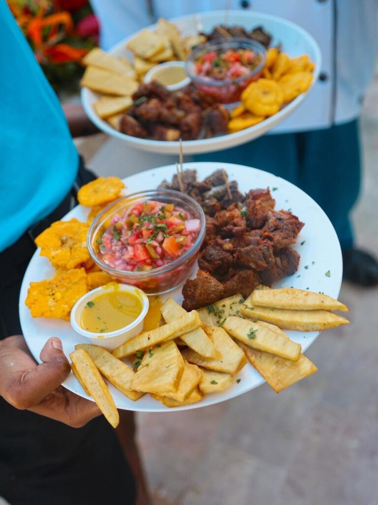A person holding a white plate of traditional Honduran food in Roatan with fries, fried meat, fresh salsa, plantains, and yellow dipping sauce with a blurred background of an outdoor setting in Honduras.