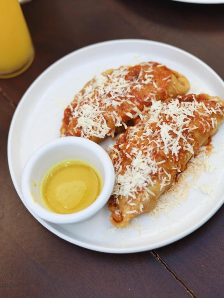 Two crispy, cheese-topped enchiladas served on a white plate with a small cup of yellow sauce and a glass of yellow beverage in the background in Roatan, Honduras.