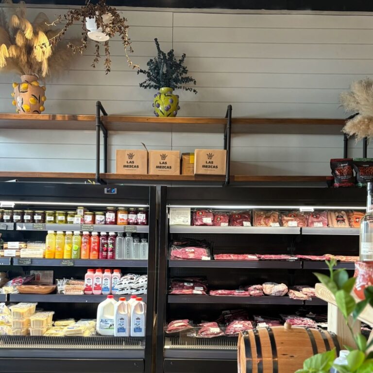 Grocery store shelf in Roatan, Honduras, displaying packaged meats, dairy products, and sauces with decorative vases and plants on the wall above.