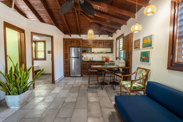 Interior view of a cozy kitchen and dining area in a Roatan home featuring wooden ceilings and modern appliances. The space includes a stainless steel refrigerator, microwave, and a water dispenser, with artwork on the walls and lush plants near the windows enhancing the warm, Tropical Honduras atmosphere.