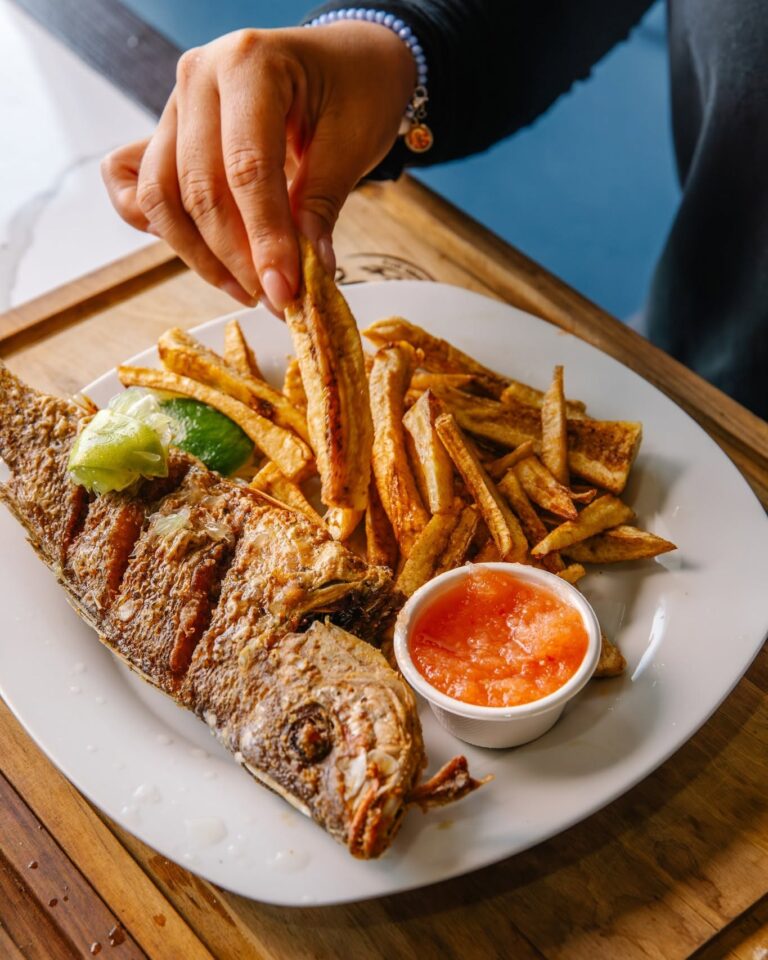 A hand holding fried fish with golden French fries, lime, and a small cup of red dipping sauce on a white plate setting in Roatan, Honduras.