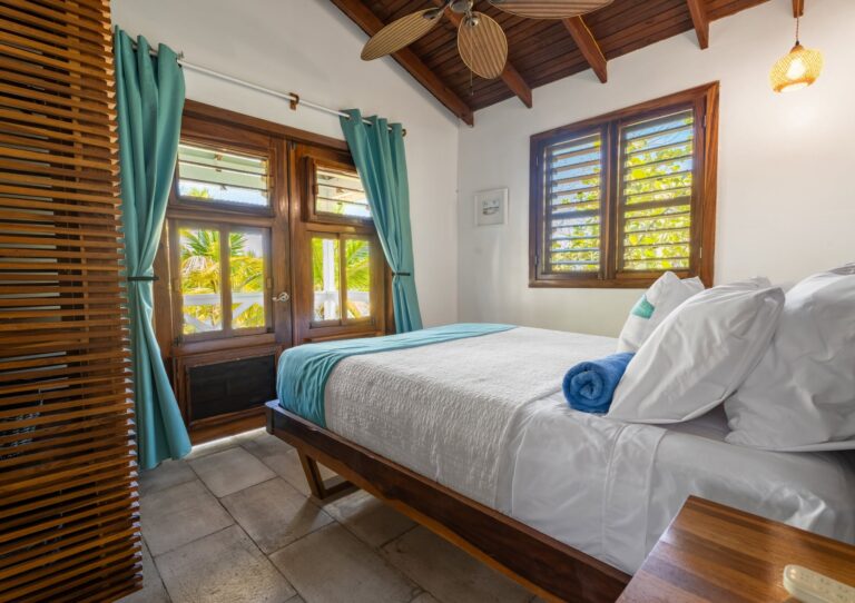 A cozy bedroom with a wooden ceiling and large windows overlooking lush green tropical plants on Roatan, Honduras, featuring a neatly made bed with white linens and turquoise accents.