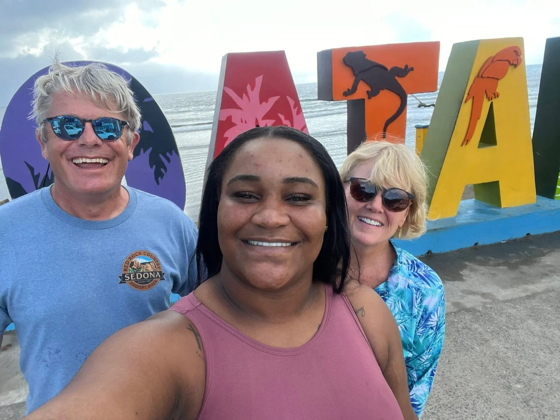 A group of smiling tourists taking a selfie in front of colorful letters spelling 'ROATAN' on the beach in Roatan, Bay Islands, Honduras, with the ocean and cloudy sky in the background.