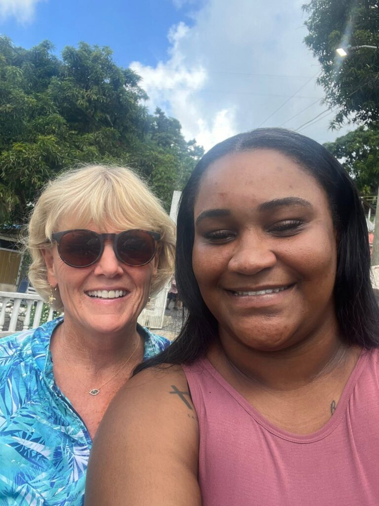 Two women, one with blonde hair and sunglasses wearing a blue patterned shirt, and the other with dark hair wearing a pink sleeveless top, smiling outdoors in Roatan, Honduras with green trees and partly cloudy sky in the background.