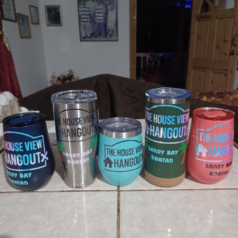 A collection of colorful tumblers with 'The House View Hangout' logo and text, placed on a white tiled surface in a cozy indoor setting in Roatan, Honduras.